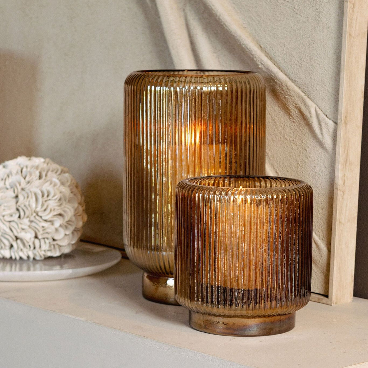 Set Van 3 Shalea Windlichten Amber Ribbelglas van PTMD, twee windlichten met brandende kaarsen op een sokkel tegen een lichte muur